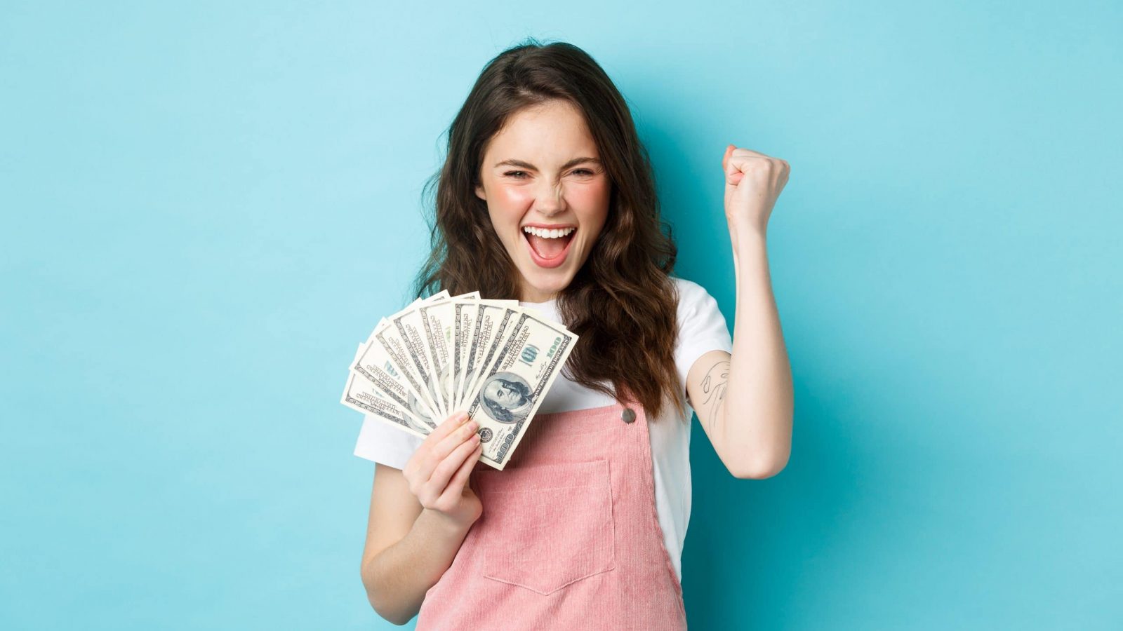Lucky young woman looks excited, shouting from satisfaction and triumph, winning money, holding dollar bills and making fist pump, standing over blue background.